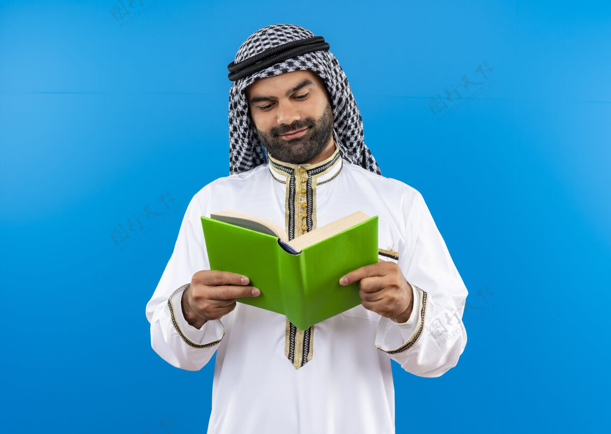 阿拉伯语身着传统服饰的阿拉伯商人手持一本打开的书 面带微笑地站在蓝色的墙上看着它看站商务人士
