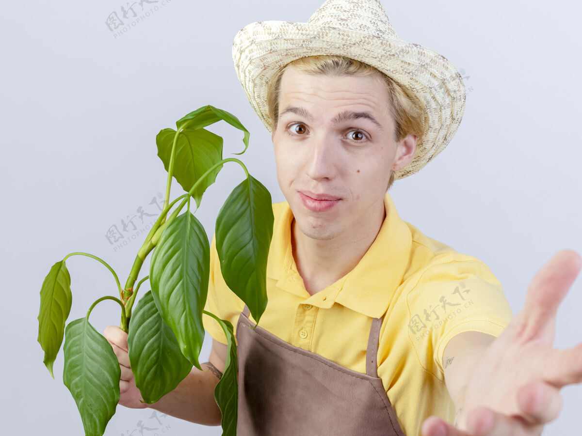 白色年轻的园丁穿着连体衣 戴着帽子 手里拿着植物 站在白色的背景上微笑着看着镜头植物抱微笑