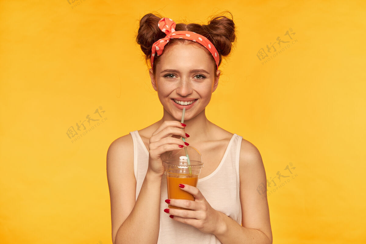 孤独年轻女士 漂亮的姜黄色女人 带着两个发髻和发圈 看起来很开心穿着白衬衫 拿着健康的冰沙微笑着看着隔离在黄色的墙上快乐牙齿红发