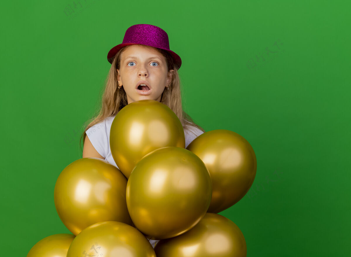 气球戴着节日礼帽的漂亮小女孩和一群巴隆人站在一边惊讶地看着 绿色背景下的生日派对概念女孩派对绿色