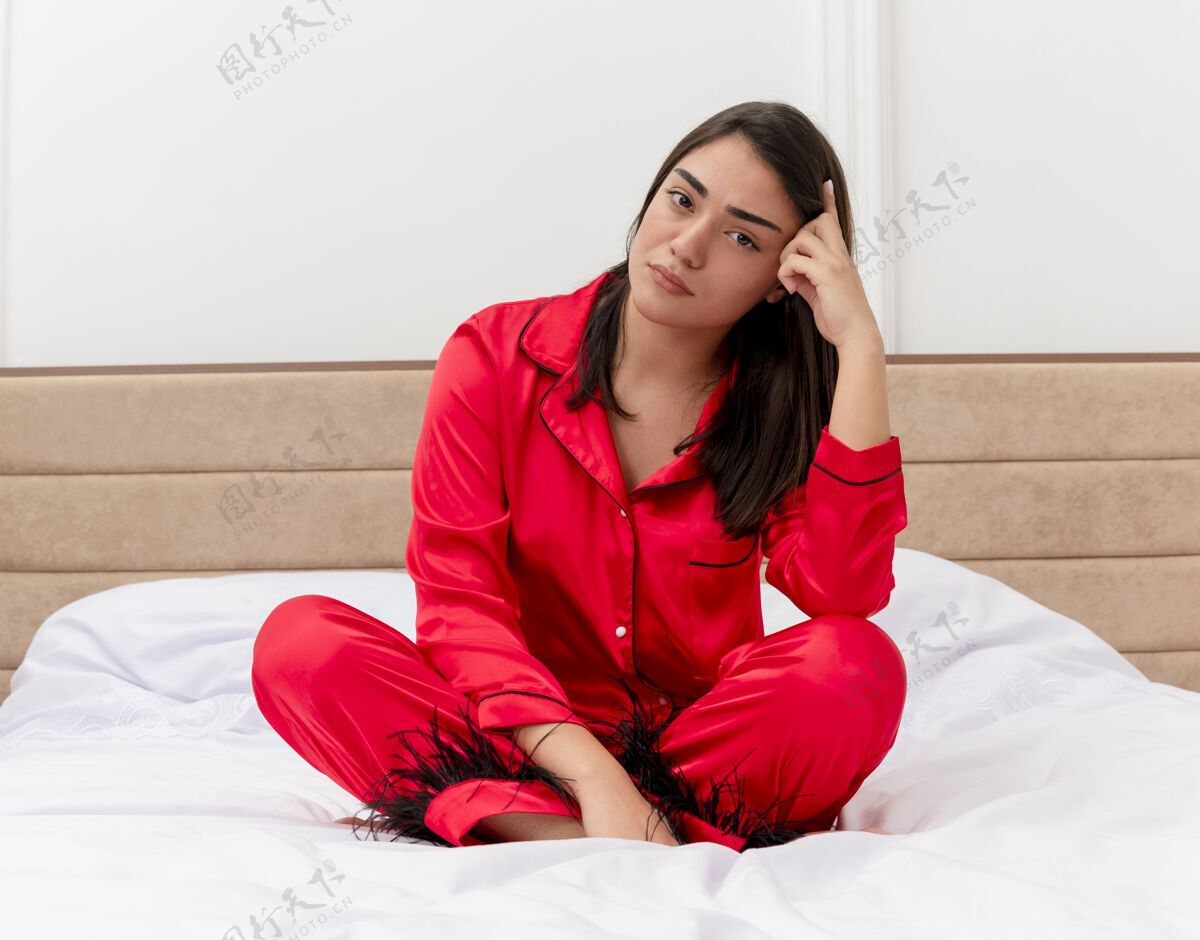 年轻穿着红色睡衣的年轻漂亮女人坐在床上看着摄像机 在卧室的室内灯光背景下有一张严肃的脸卧室睡衣坐着