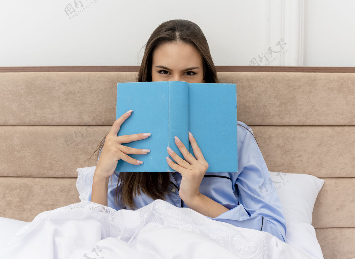 漂亮穿着蓝色睡衣的年轻美女坐在床上 书背上的脸在卧室里的灯光背景下偷看年轻床书
