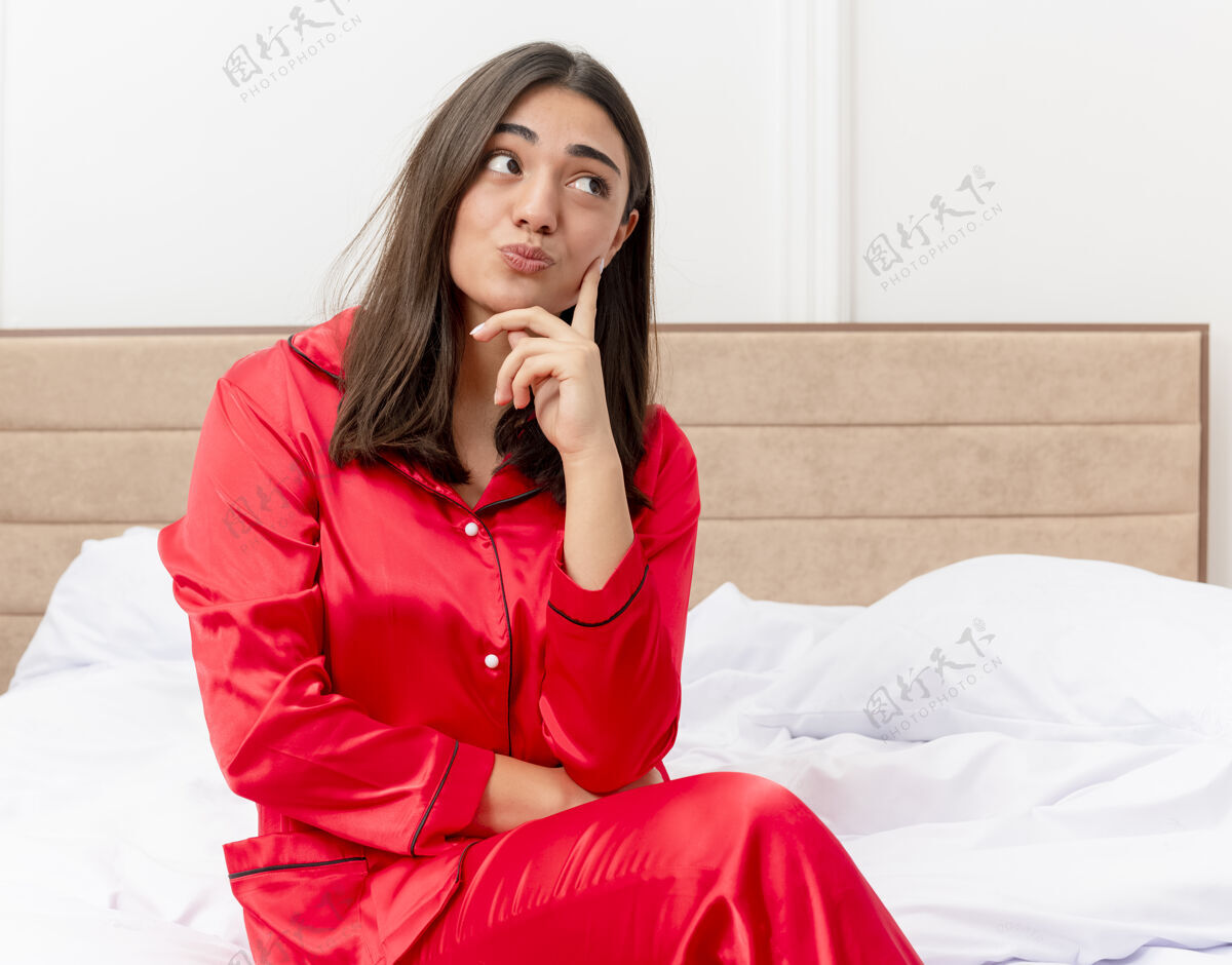 思考穿着红色睡衣的年轻美女坐在床上仰望 脸上挂着沉思的表情 在卧室内部的灯光背景下思考表情美丽看