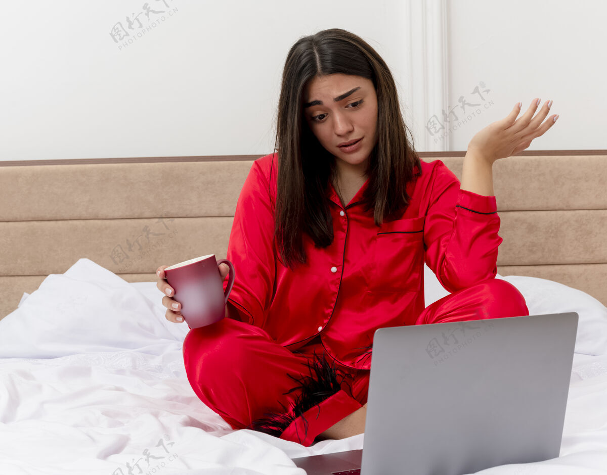 看着穿着红色睡衣的年轻美女坐在床上 拿着笔记本电脑和一杯咖啡 在浅色背景下的卧室里显得困惑和失望失望卧室红色