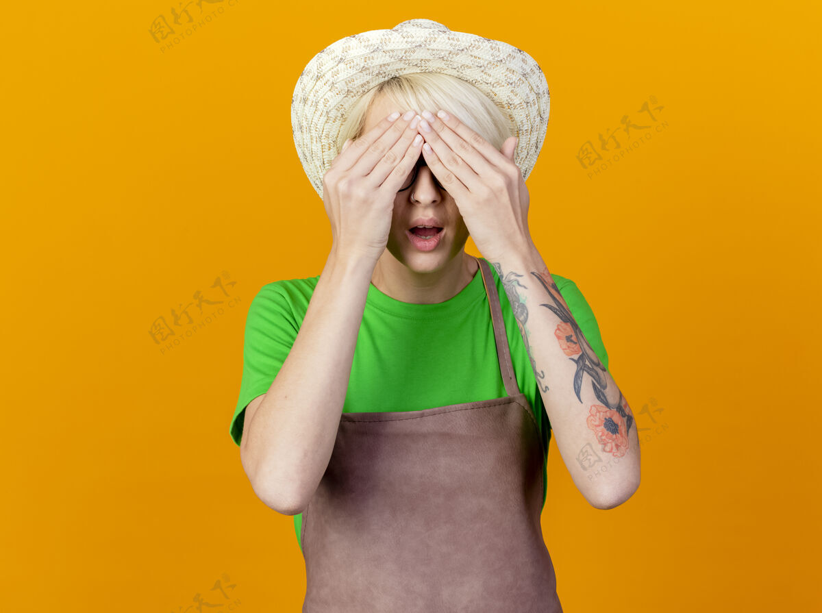 短裤围裙短发 戴帽子 眼睛蒙着的年轻园丁女士站在橙色背景下惊讶不已花园橙色头发
