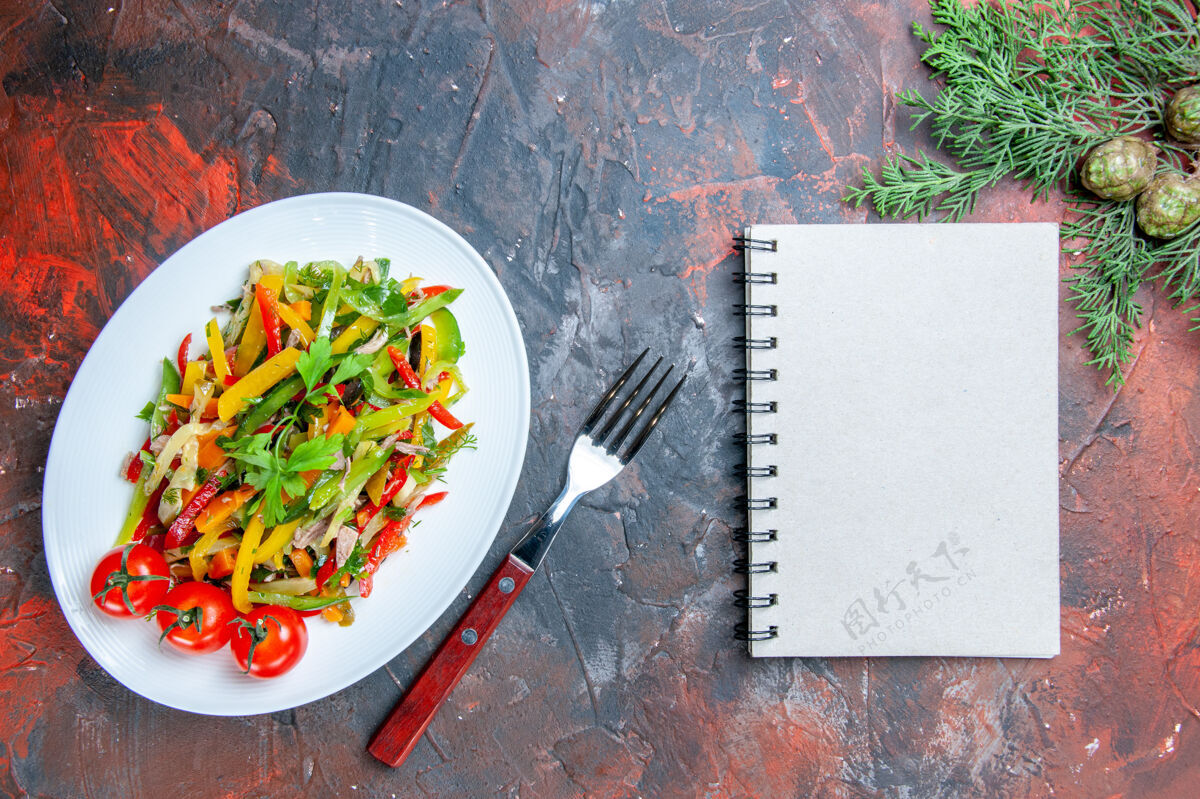 菜肴俯瞰蔬菜沙拉在椭圆形的盘子叉笔记本松枝在暗红色的桌子上沙拉盘子餐厅