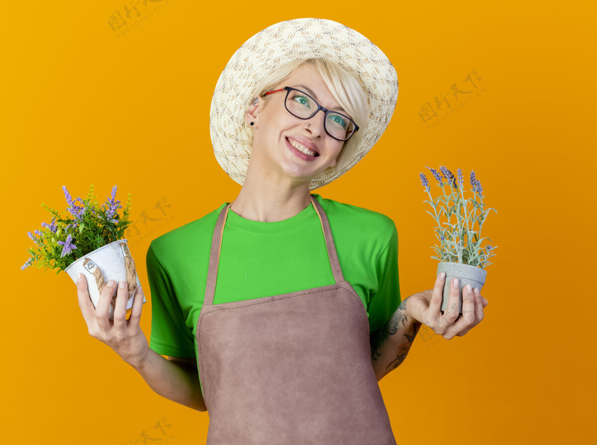 帽子围裙里留着短发 戴着帽子 手里拿着盆栽植物的年轻园丁站在橙色的背景上 面带微笑女人微笑围裙