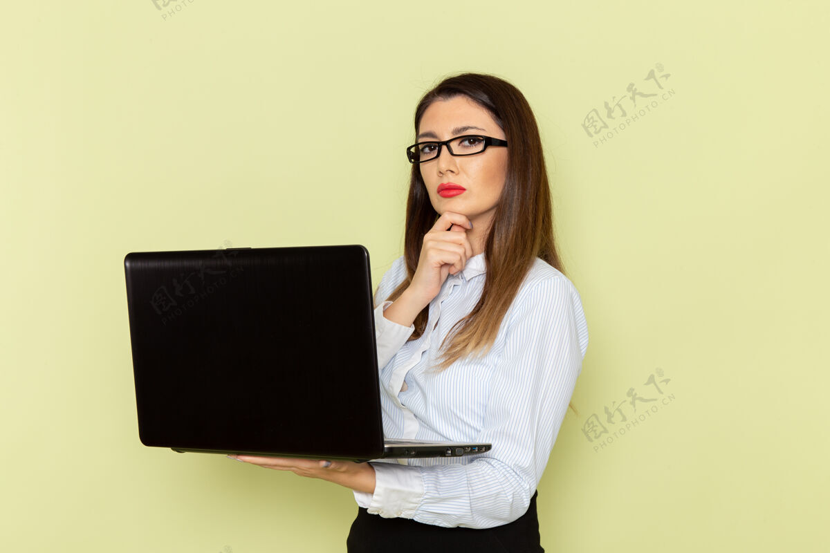 办公室穿着白衬衫和黑裙子的女上班族在浅绿色墙上使用笔记本电脑的正视图商务衬衫电脑