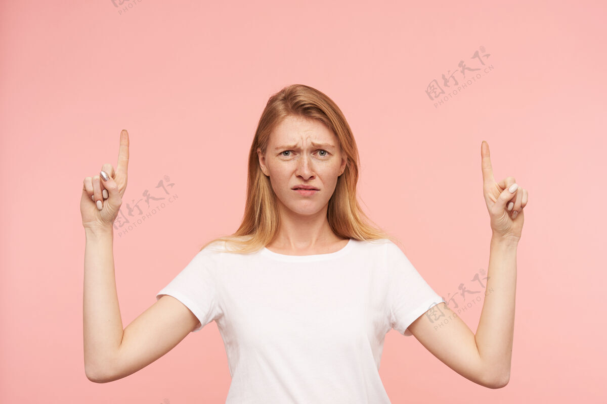 肖像困惑的年轻漂亮的红发女性 随意的发型出现在食指上 站在粉色背景下困惑地皱起眉头手指头发白色
