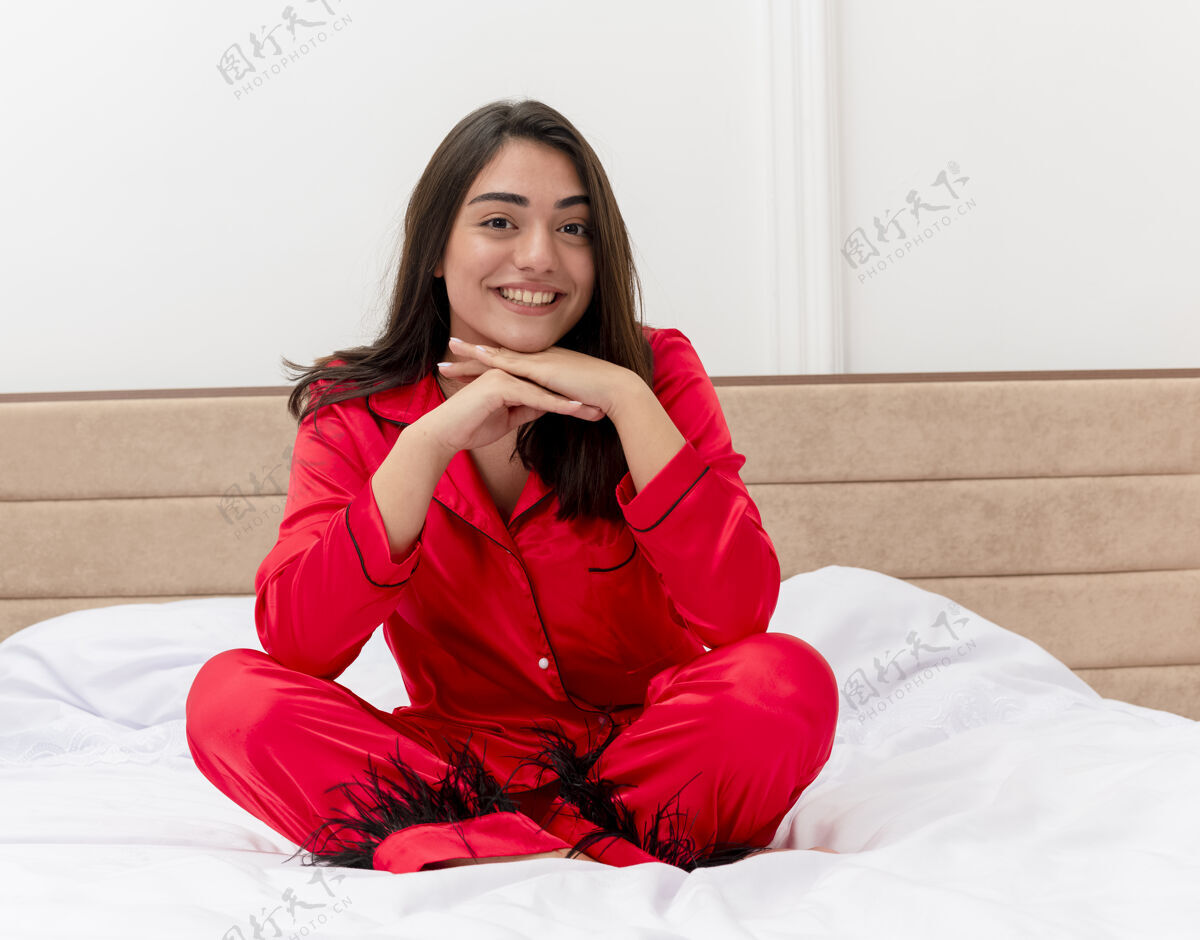 红色穿着红色睡衣的年轻漂亮女人坐在床上微笑着 脸上洋溢着幸福的表情 在卧室内部的灯光背景下把头靠在手掌上对着摄像机摆姿势头瘦漂亮