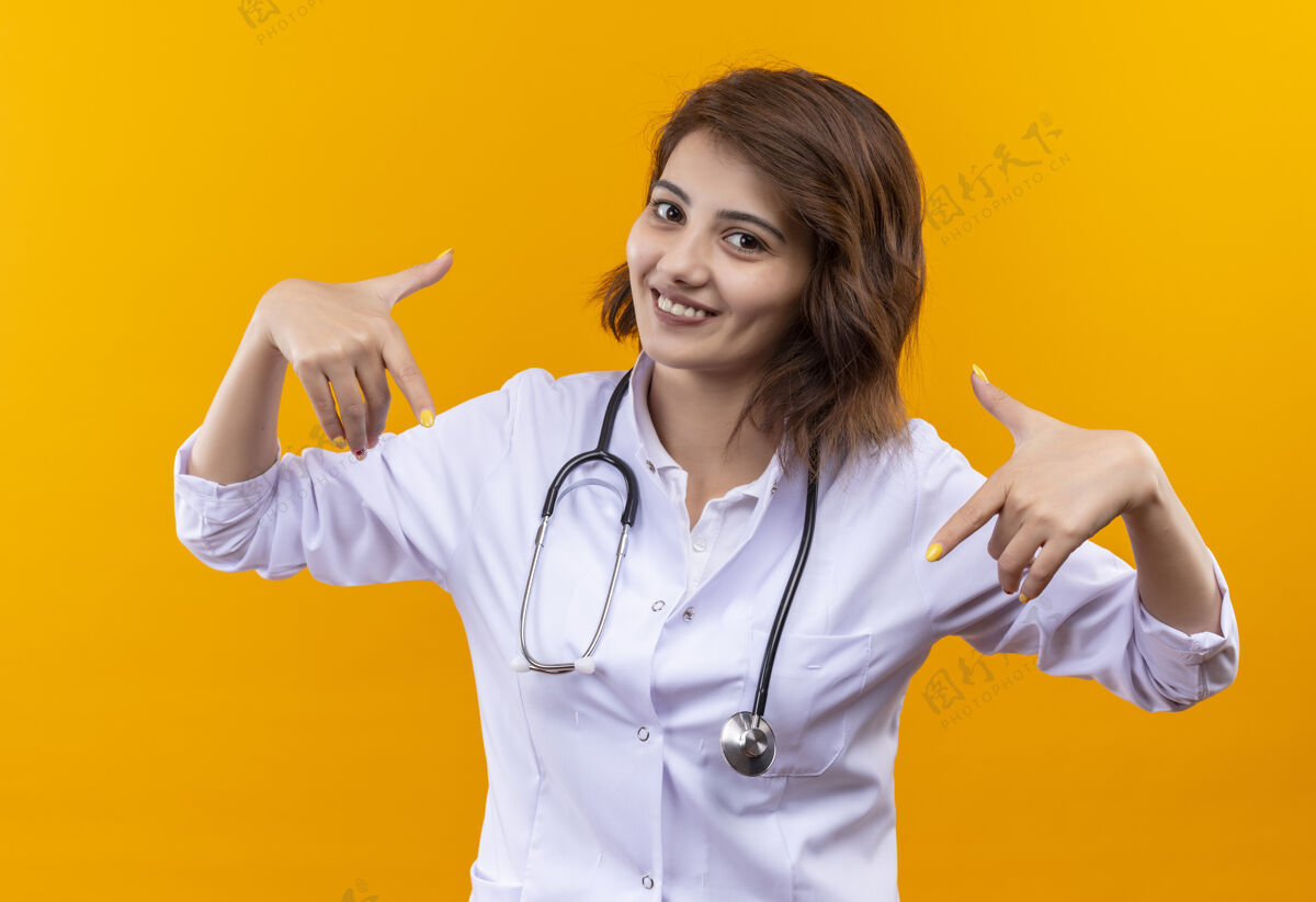 阳性穿着白大褂的年轻女医生拿着听诊器快乐而积极地指着自己听诊器快乐医生
