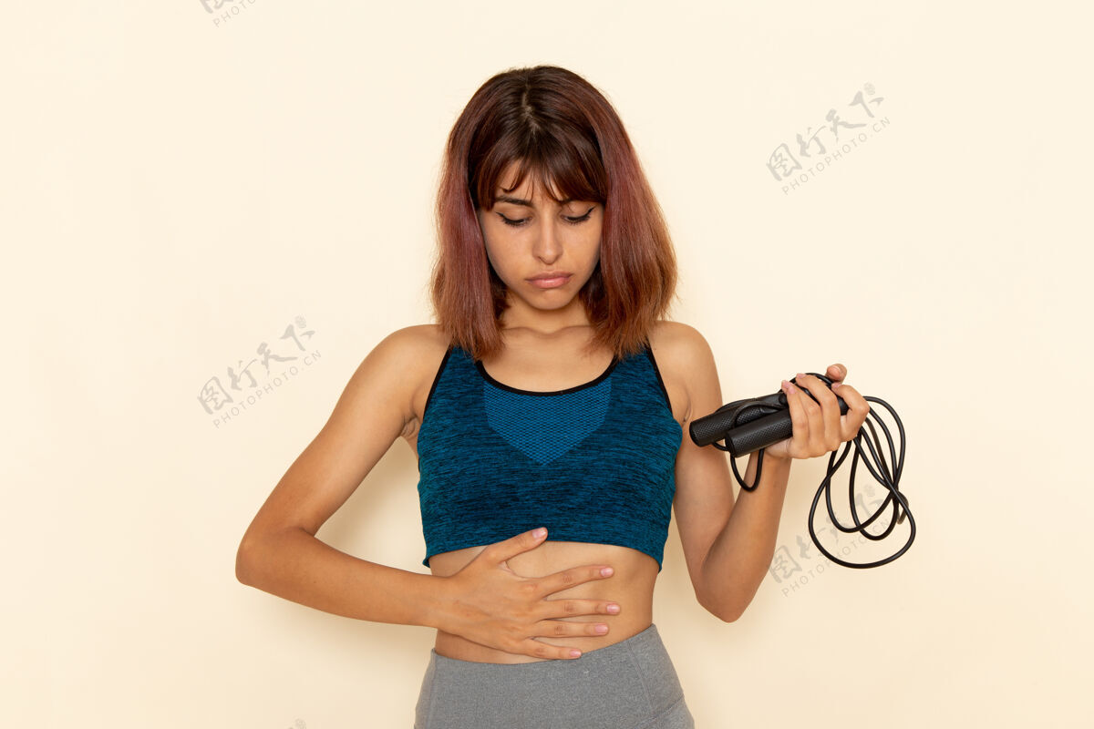 绳子前视图中的年轻女性身体健康 穿着蓝色衬衫 手持跳绳站在浅白的墙上运动身体前面