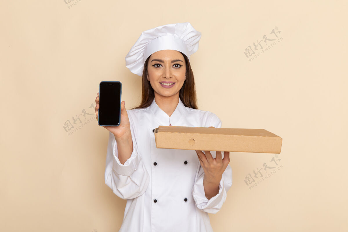 套装身穿白色厨师服的年轻女厨师的正视图 在浅白的墙上拿着电话和食物盒烹饪电话工作