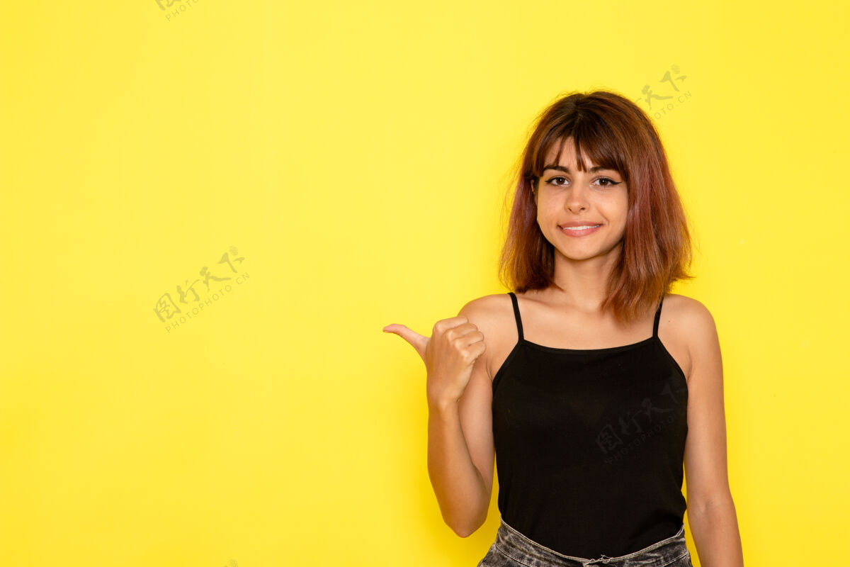 性感身穿黑色衬衫和灰色牛仔裤的年轻女性在黄色墙壁上微笑和摆姿势的正面视图模特姿势漂亮