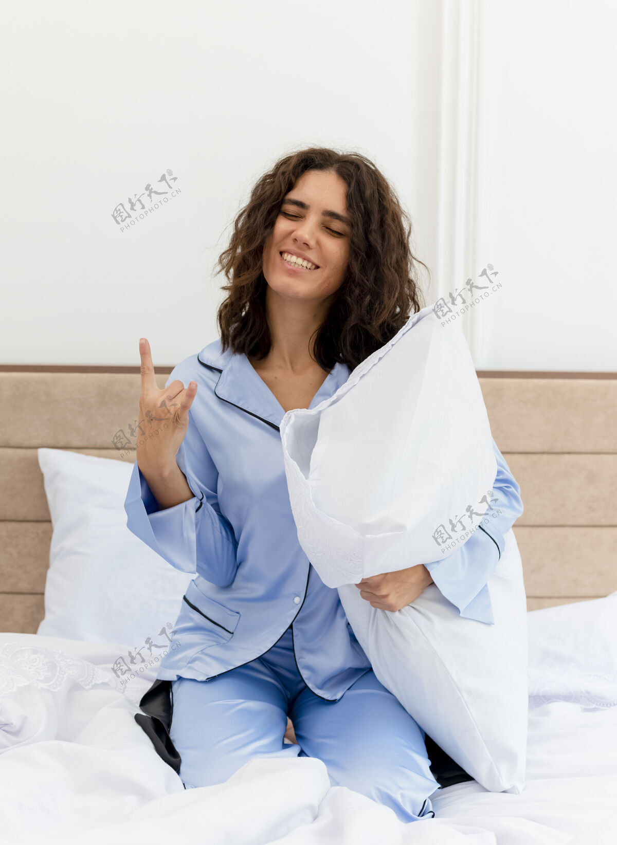 女人穿着蓝色睡衣的年轻漂亮女人坐在床上 带着枕头 快乐而积极的微笑 在卧室内部的灯光背景下展现摇滚乐的象征枕头摇滚展示