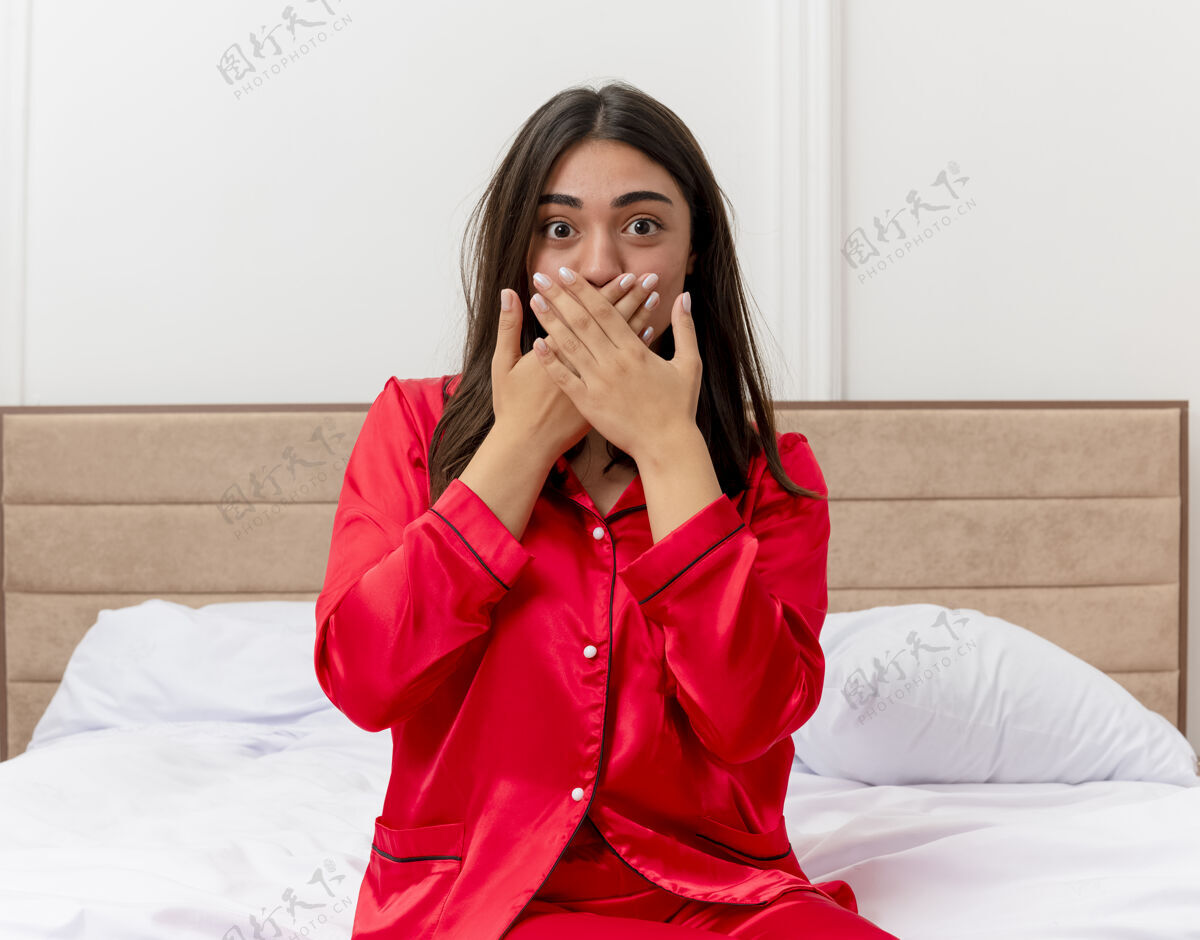 卧室穿着红色睡衣的年轻美女坐在床上看着摄像机 惊讶地用手捂住嘴巴 在卧室的室内灯光背景下嘴红色相机