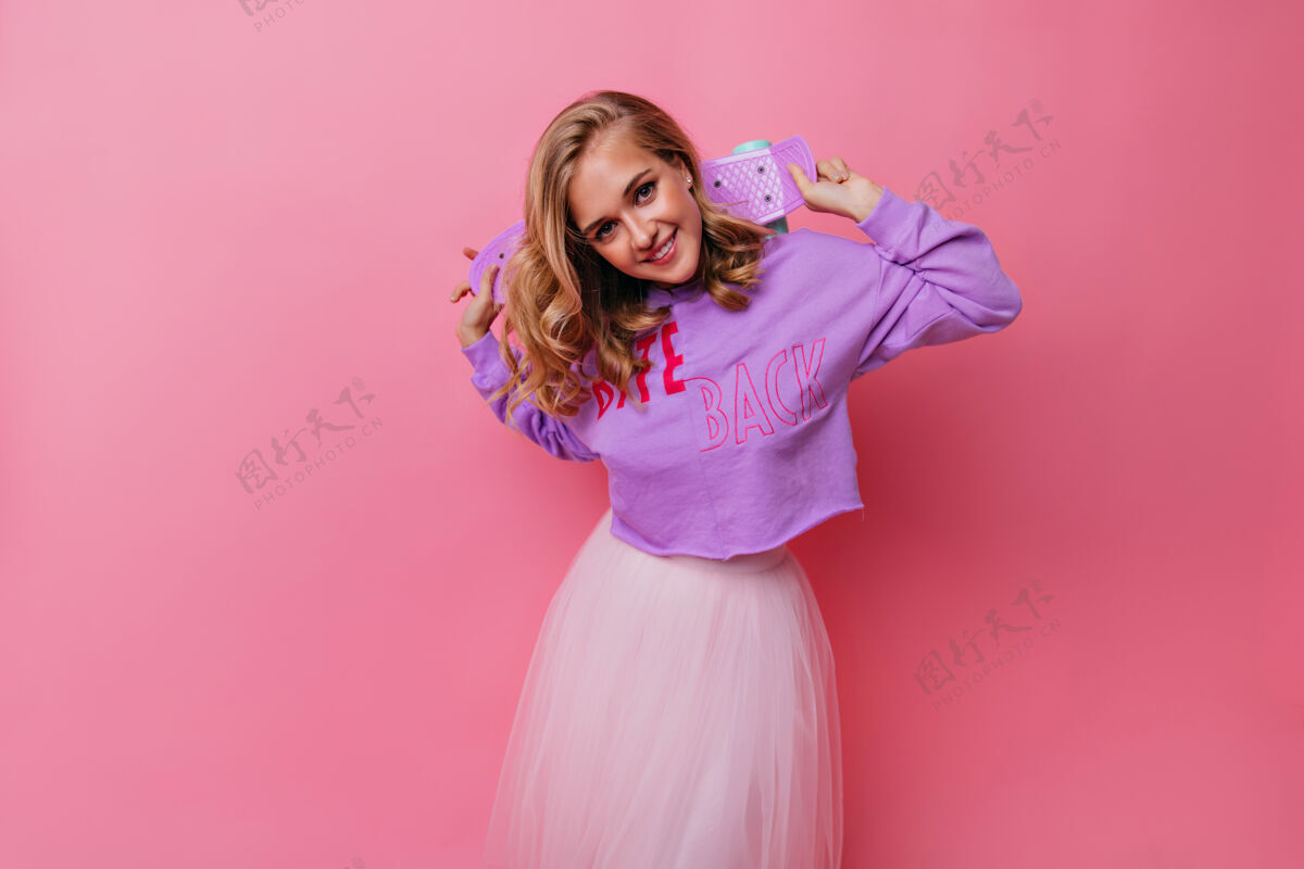 配件穿着时髦的紫色衬衫拿着滑板的壮观的女人欣喜若狂的卷曲金发女孩的室内肖像放松滑板装置