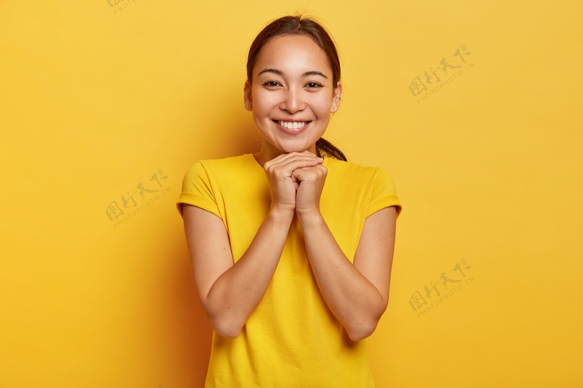 积极照片中富有魅力的亚洲女性双手合十 面带微笑 表情可爱 深色头发梳成马尾辫 穿着鲜艳的黄色t恤 在令人敬畏的同伴中娱乐学生请休闲