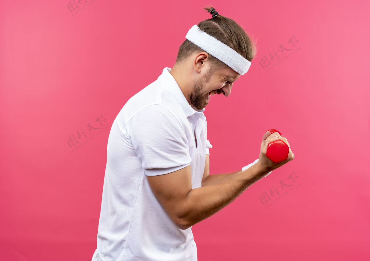 男人戴着头带和腕带的瘦骨嶙峋的年轻帅气运动型男子站在侧视图中 举着哑铃 闭着眼睛孤立在粉红色的空间里紧张运动头带