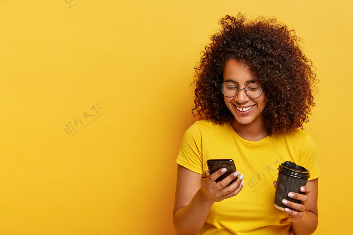 眼镜快乐的卷发少女 手持现代手机 外卖咖啡 通过在线应用订购出租车 输入短信 穿着黄色衣服人 现代生活方式和技术千禧年高兴通讯