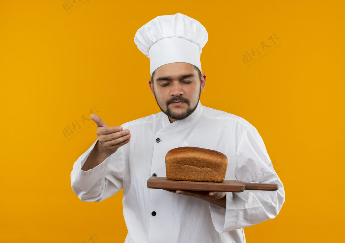 厨师身着厨师制服的年轻男厨师手拿着放着面包的砧板 手放在空气中嗅着 闭着眼睛孤立在橙色的空间里面包关闭板