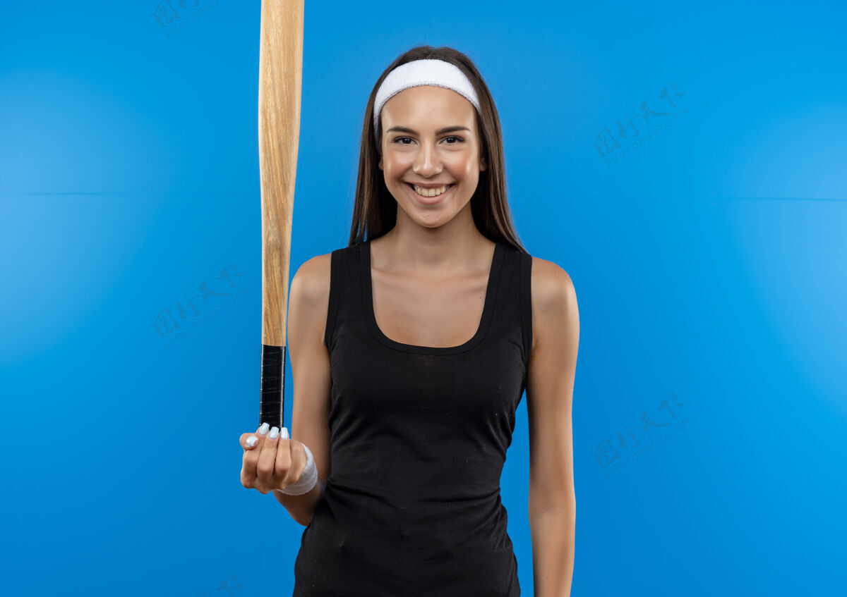 年轻微笑的年轻漂亮的运动女孩戴着头带和手环拿着棒球棒孤立在蓝色的空间微笑太空穿着