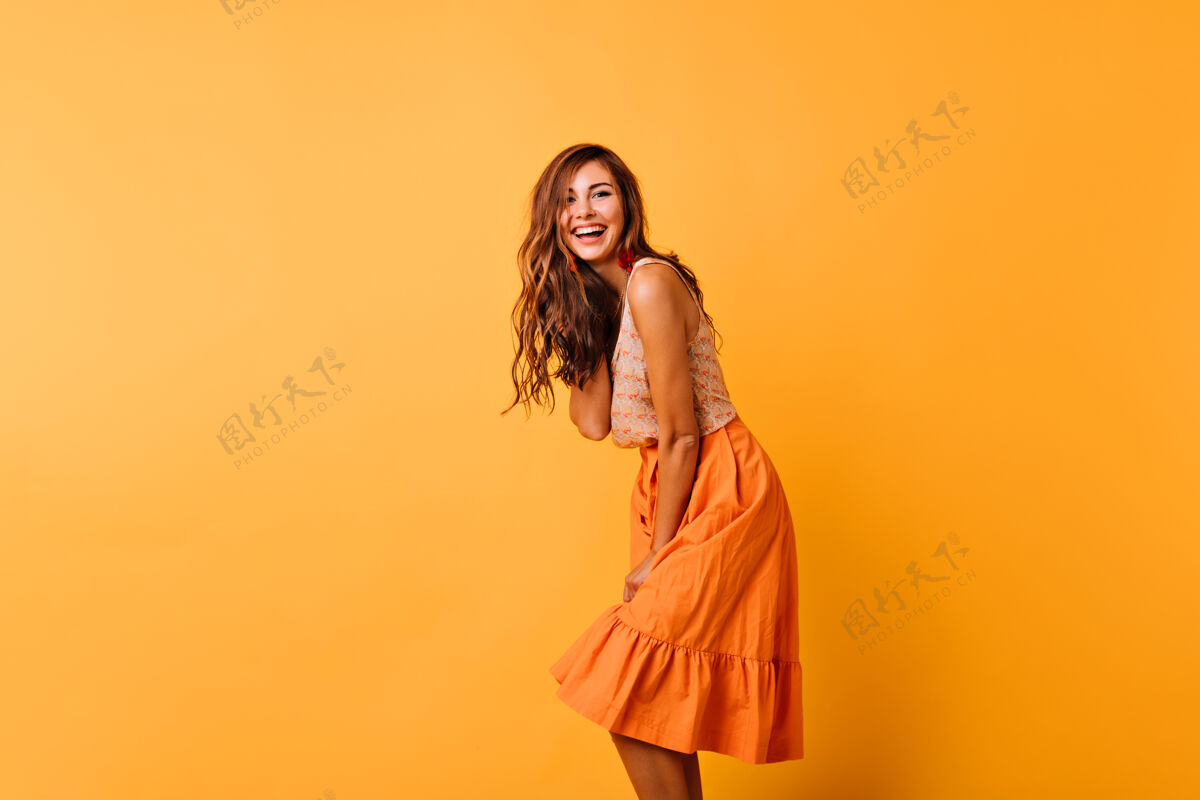 模特长发卷曲的女人 穿着鲜艳的服装 表达积极的情绪穿着橙色裙子的浪漫女模特在黄色的裙子上跳舞表情放松狂喜
