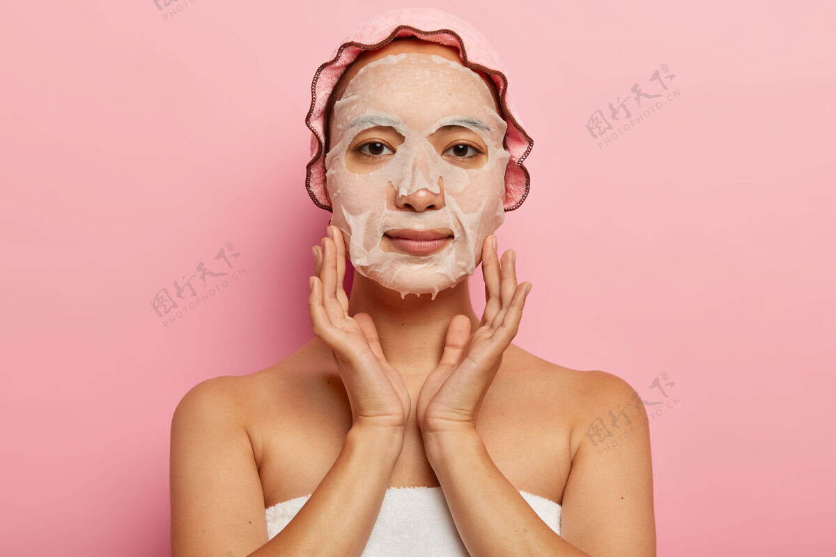 毛巾严肃的日本女性在脸上贴上滋养面膜 在皮肤上涂上保湿单品 戴上浴帽 对着粉色墙壁摆姿势女性化 美容和spa治疗理念肩膀面膜保持
