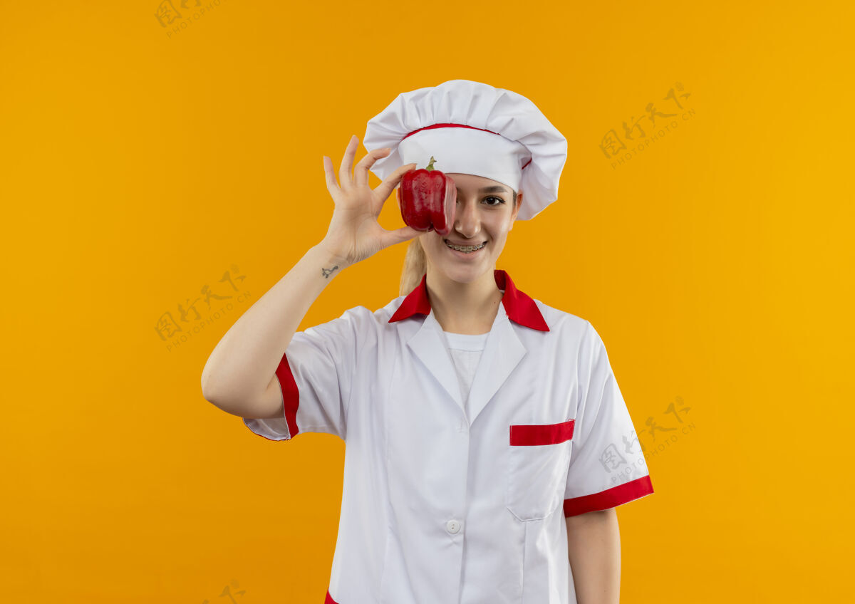 年轻微笑着的年轻漂亮厨师身着厨师制服 戴着牙套 把胡椒粉放在眼睛上 隔离在橙色的空间里眼睛制服牙科