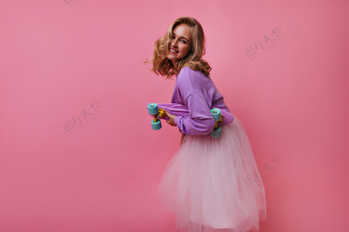 装置迷人的女模特穿着华丽的裙子和紫色衬衫与滑板合影有趣的金发女孩嬉戏的室内肖像舞蹈欢笑乐趣