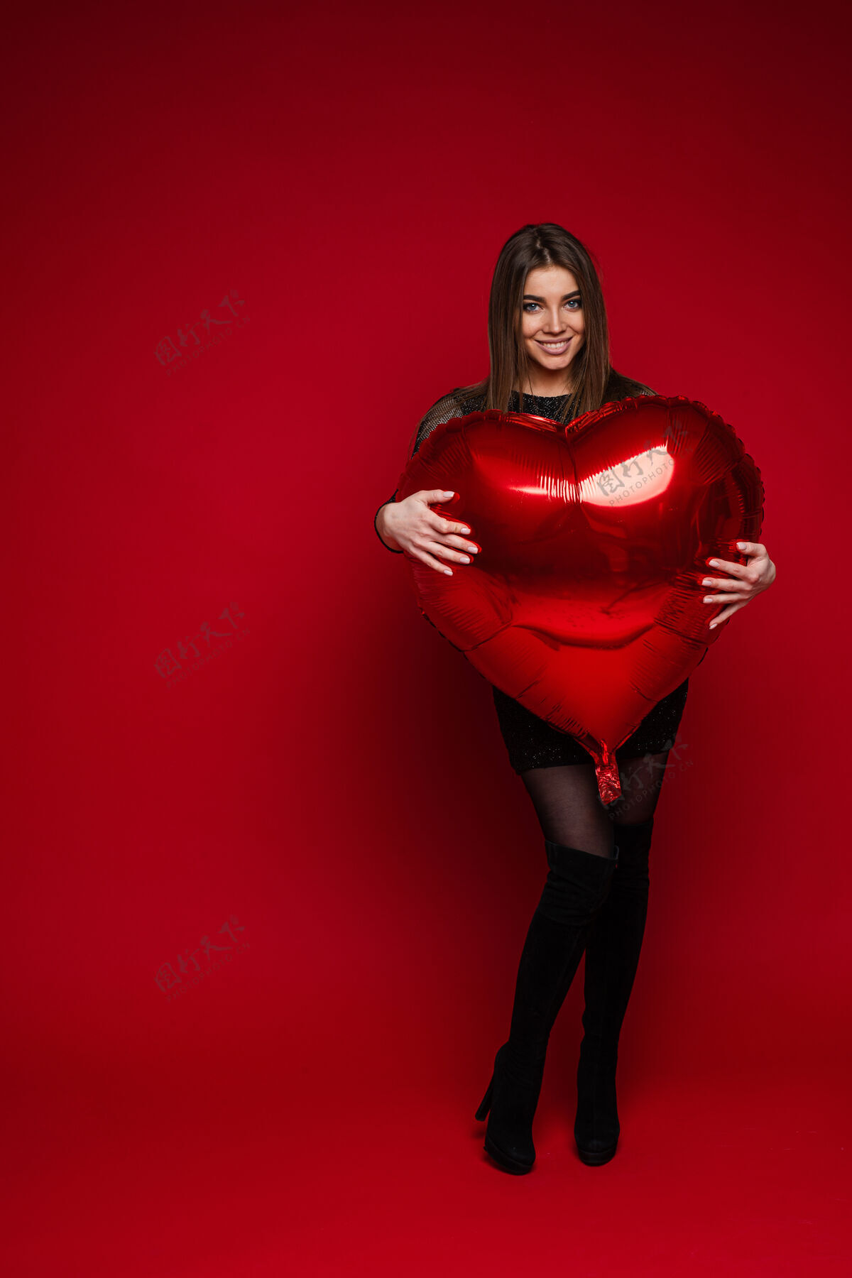 爱身着裙子和靴子的快乐黑发女孩在红色背景上拥抱红色心形气球的全长肖像圣瓦伦丁概念情人节节日2月14日