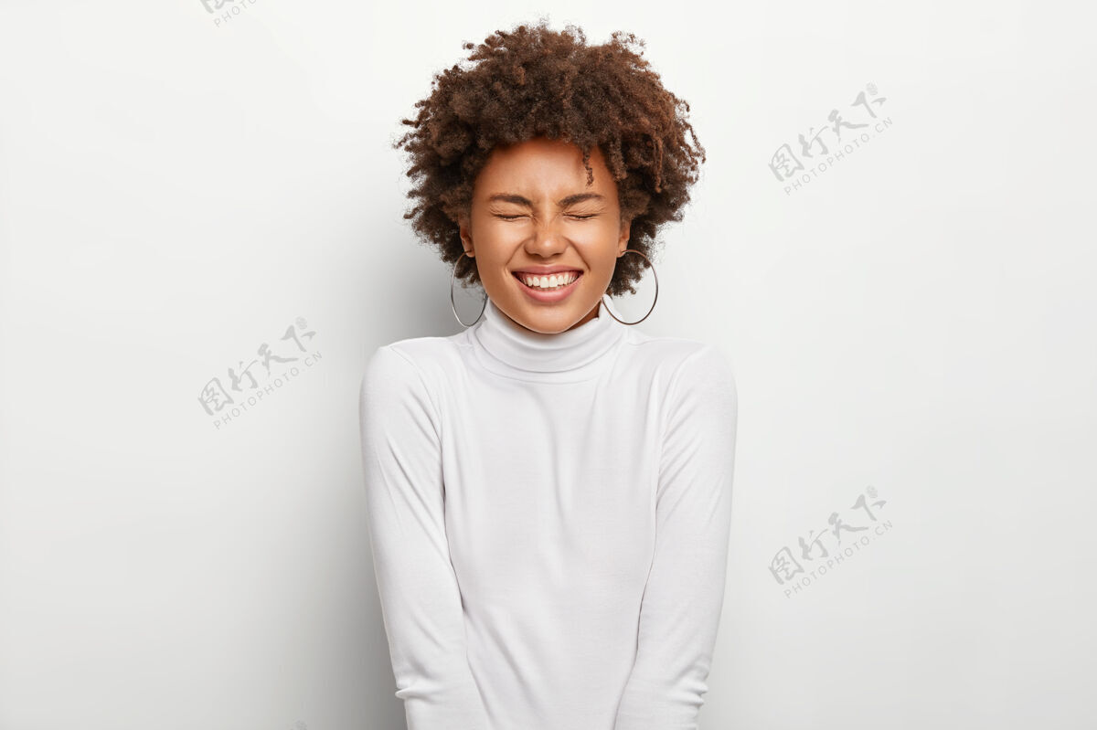 女性过度活跃的美国黑人女士积极地笑出声来 闭上眼睛 对有趣的故事微笑 表达良好的情感 穿着白色衣服 与世隔绝 留着卷发人与情感魅力欣喜若狂年轻