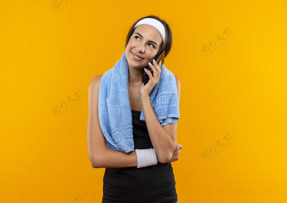 会说话带着微笑的年轻漂亮的运动女孩戴着头带和腕带 脖子上围着毛巾在橙色的空间里打电话运动穿着头带