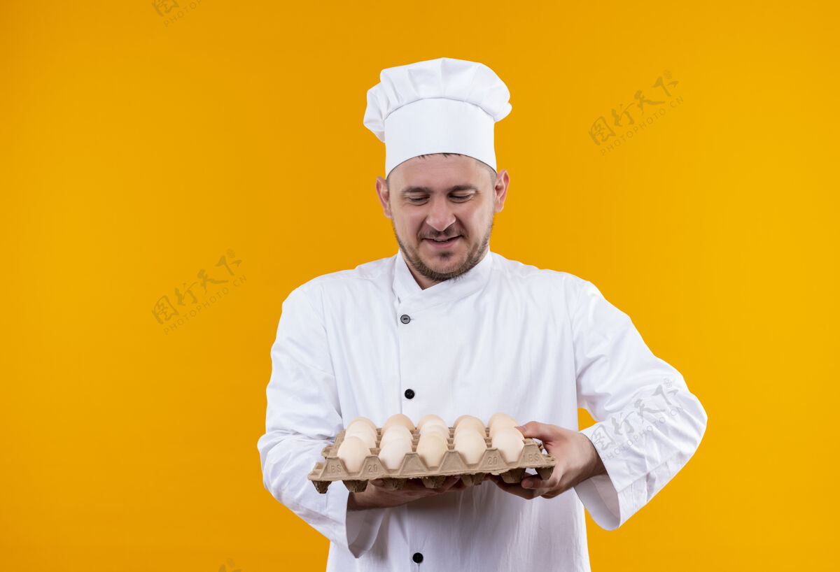 制服年轻帅气的厨师手拿厨师制服 看着孤立在橙色空间里的一箱鸡蛋年轻持有鸡蛋