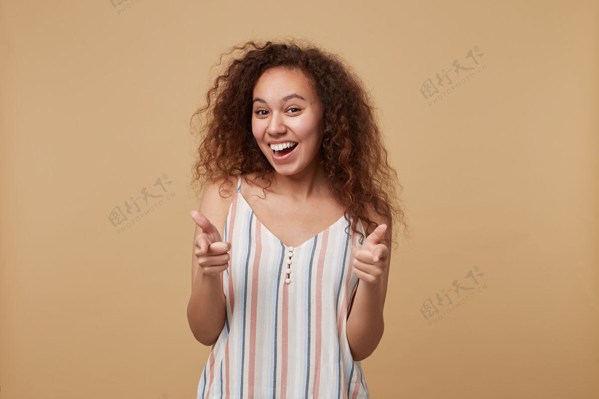 深色快乐的年轻棕色眼睛长发卷曲的女性高兴地微笑着 用食指举起她的手 孤立在米色漂亮米色肖像