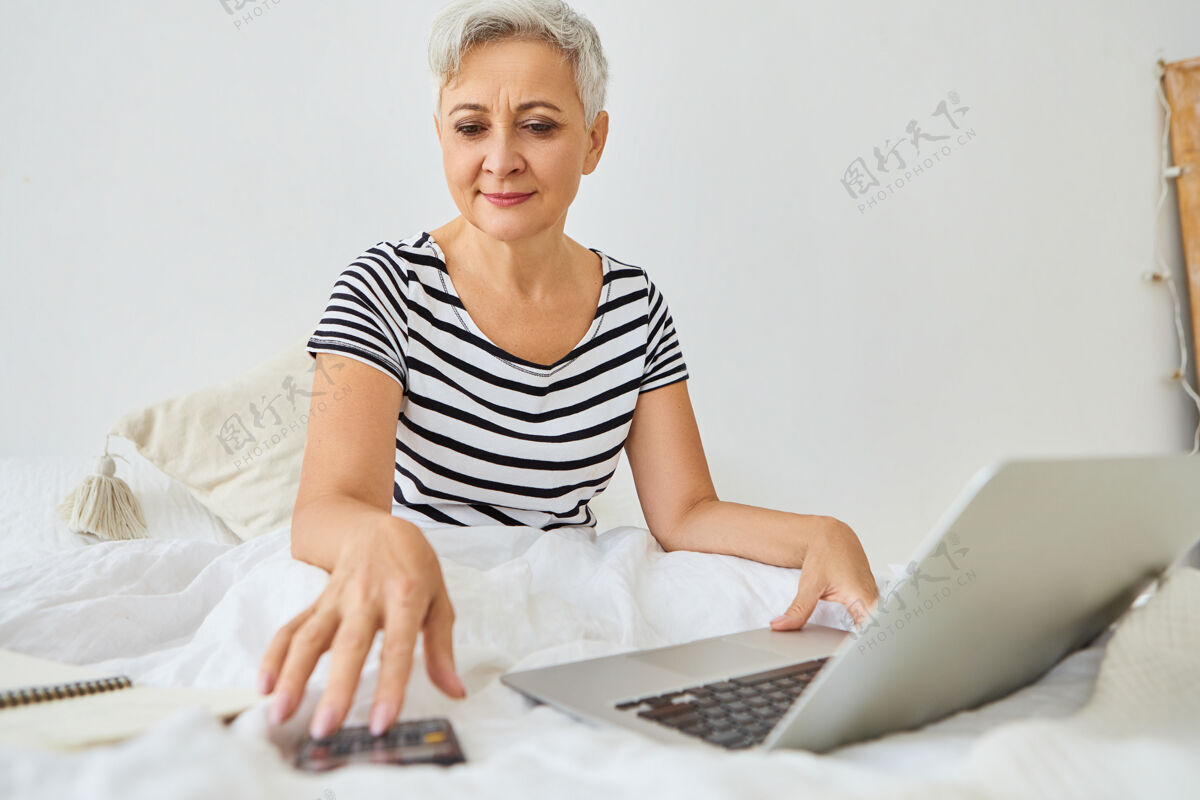 公寓迷人的白发苍苍的老商人从卧室里远程工作 坐在床上用便携式电脑 使用计算器 管理财务 有自信快乐的表情成熟遥远使用