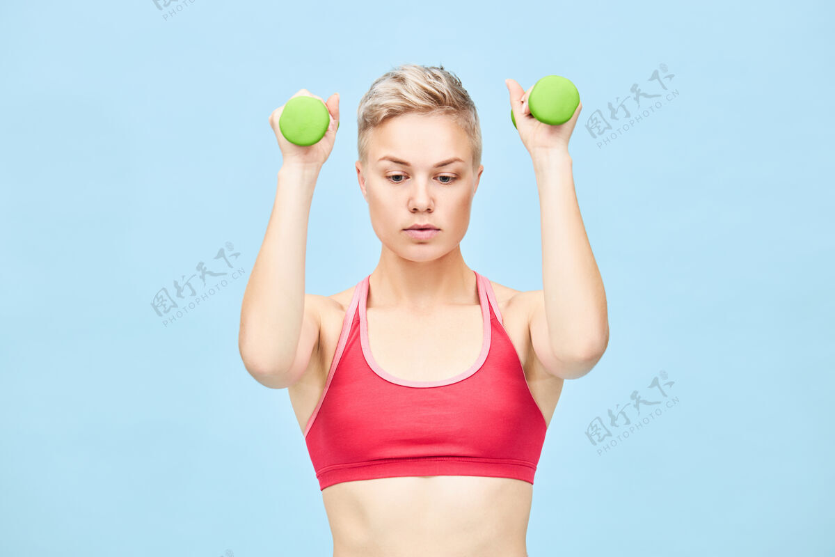 女子健身 健康和运动的概念一个年轻的欧洲金发碧眼的女人在时尚的红色上衣做二头肌卷发 举起两个绿色的哑铃 建立手臂肌肉 有重点的样子孤立的镜头肌肉健美健身