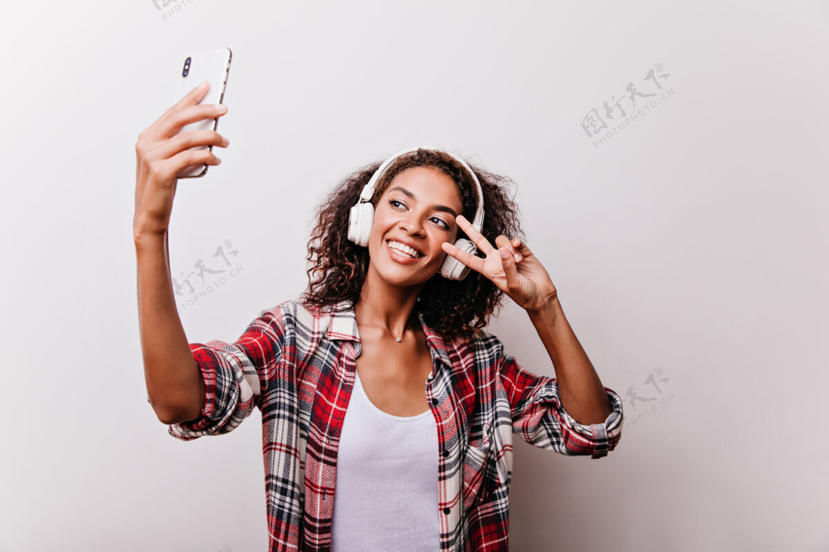 女士迷人的黑发女孩与和平标志自拍迷人的年轻女子戴着白色耳机手持智能手机年轻现代模特