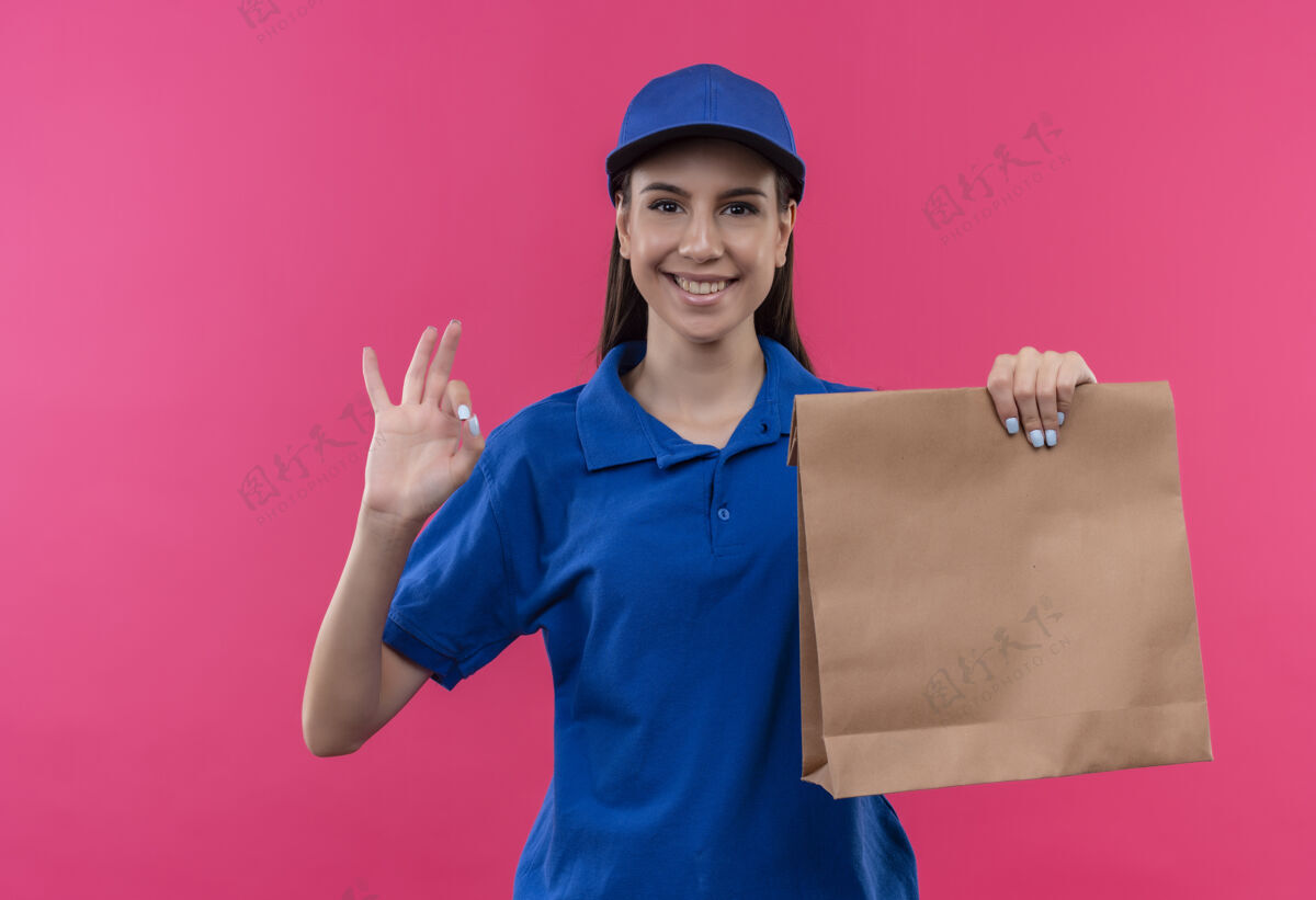 纸身穿蓝色制服 头戴鸭舌帽的年轻送货员拿着纸包看着摄像机 兴高采烈地微笑着 露出“ok”的手势帽子包装好