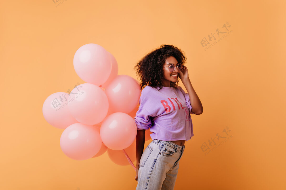 可爱戴着眼镜的时髦卷发女人手里拿着粉色的氦气球 笑着高兴的非洲生日女孩孤立在橙色上惊喜表情生日