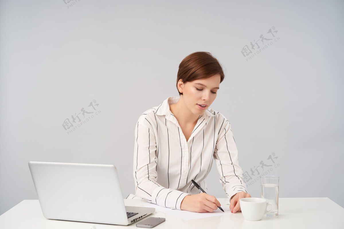 陶瓷忙碌的年轻漂亮短发黑发女性 随意的发型 在重要会议前拿着笔做笔记 穿着白色条纹衬衫钢笔深色玻璃