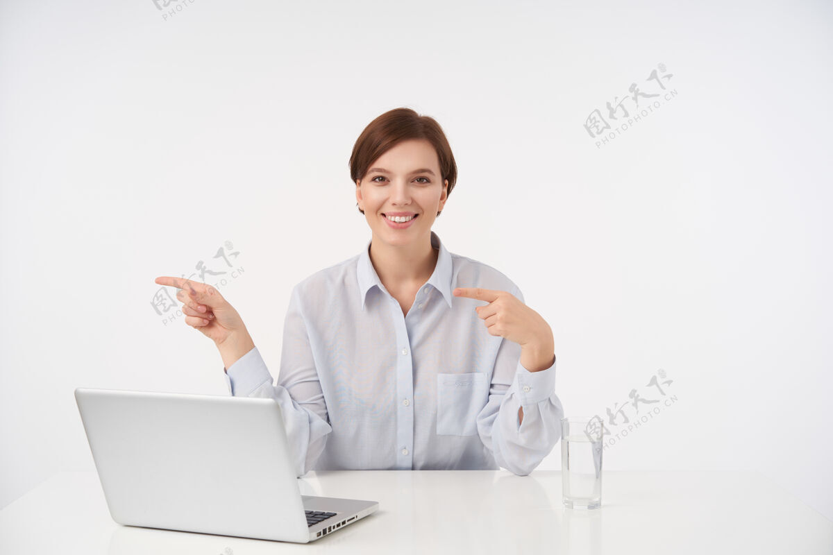 休闲快乐的年轻漂亮的短发黑发女人在办公室里拿着她的笔记本电脑 面带微笑 正眼看着 坐在白色的椅子上用食指指着旁边现代员工女人
