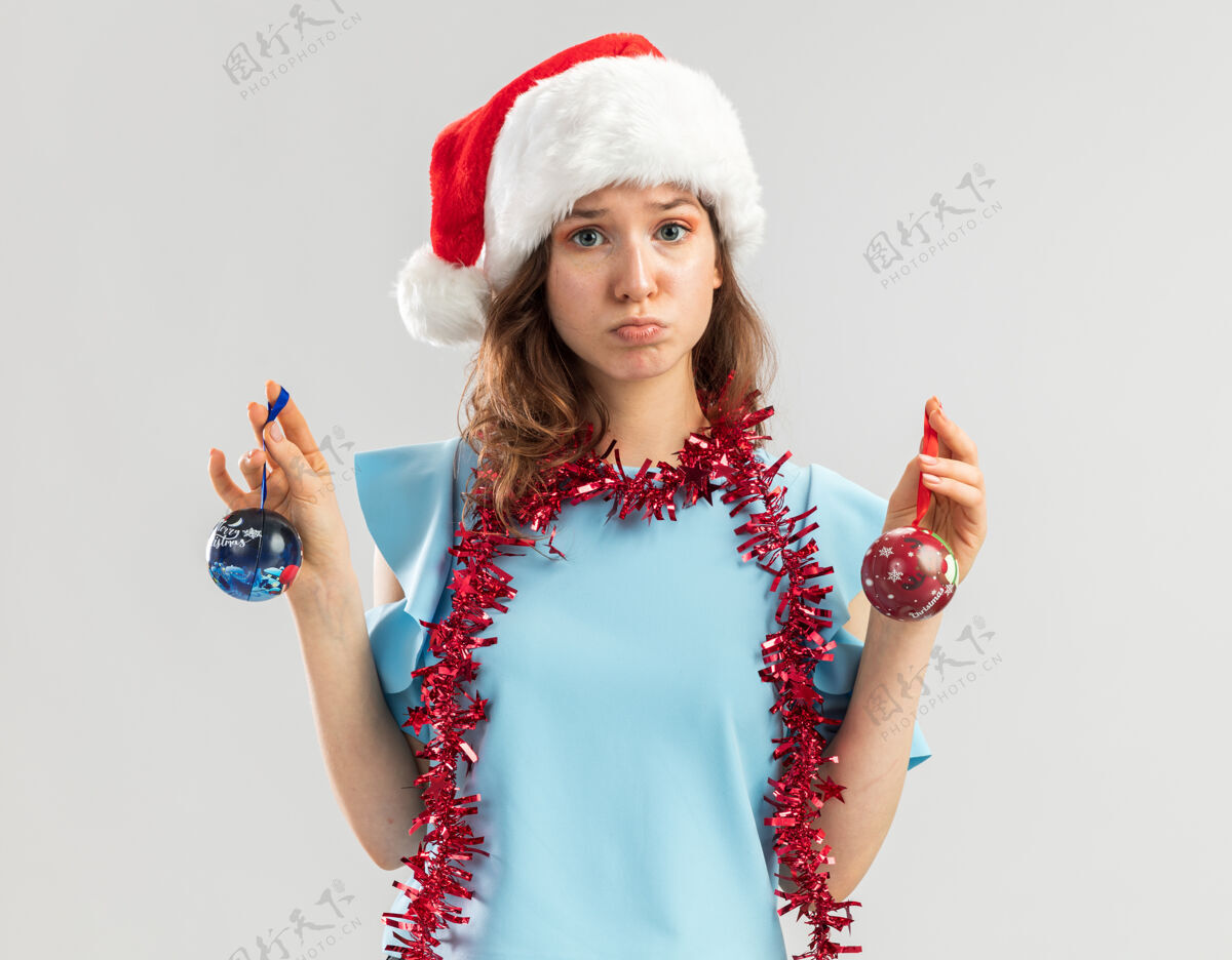 年轻身穿蓝色上衣 头戴圣诞帽 脖子上戴着金属箔的年轻女子拿着圣诞球 看上去既困惑又不高兴金属片年圣诞