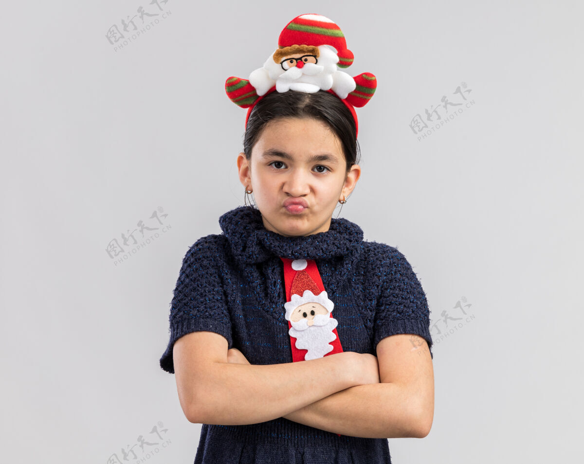 年愤愤不平的小女孩穿着针织连衣裙 头上系着一条红色领带 戴着滑稽的圣诞戒指 两臂交叉 满脸怒容小编织新