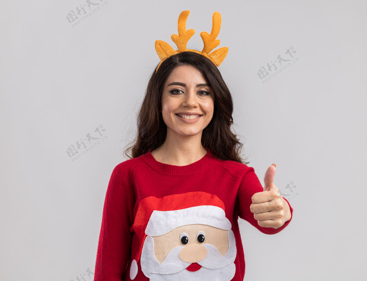 拇指微笑的年轻漂亮女孩戴着驯鹿鹿角头带和圣诞老人毛衣 看起来大拇指向上圣诞快乐驯鹿展示