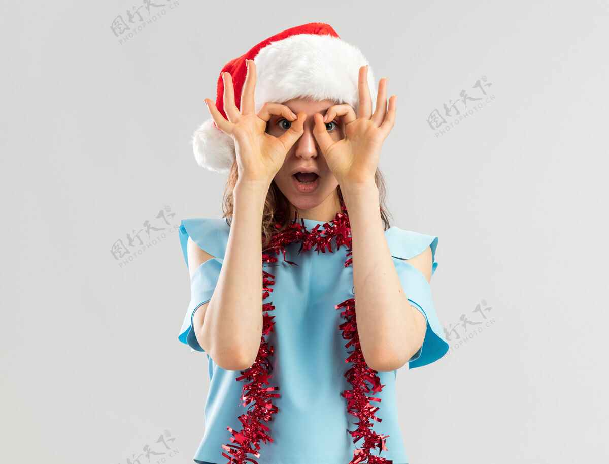 惊讶身穿蓝色上衣 戴着圣诞帽 脖子上戴着金属丝的年轻女子透过手指做着双目手势 惊讶不已通过脖子存在
