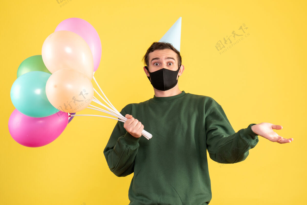 伪装正面图：戴着派对帽 站在黄色地面上的彩色气球的年轻人黑色喜剧演员五颜六色