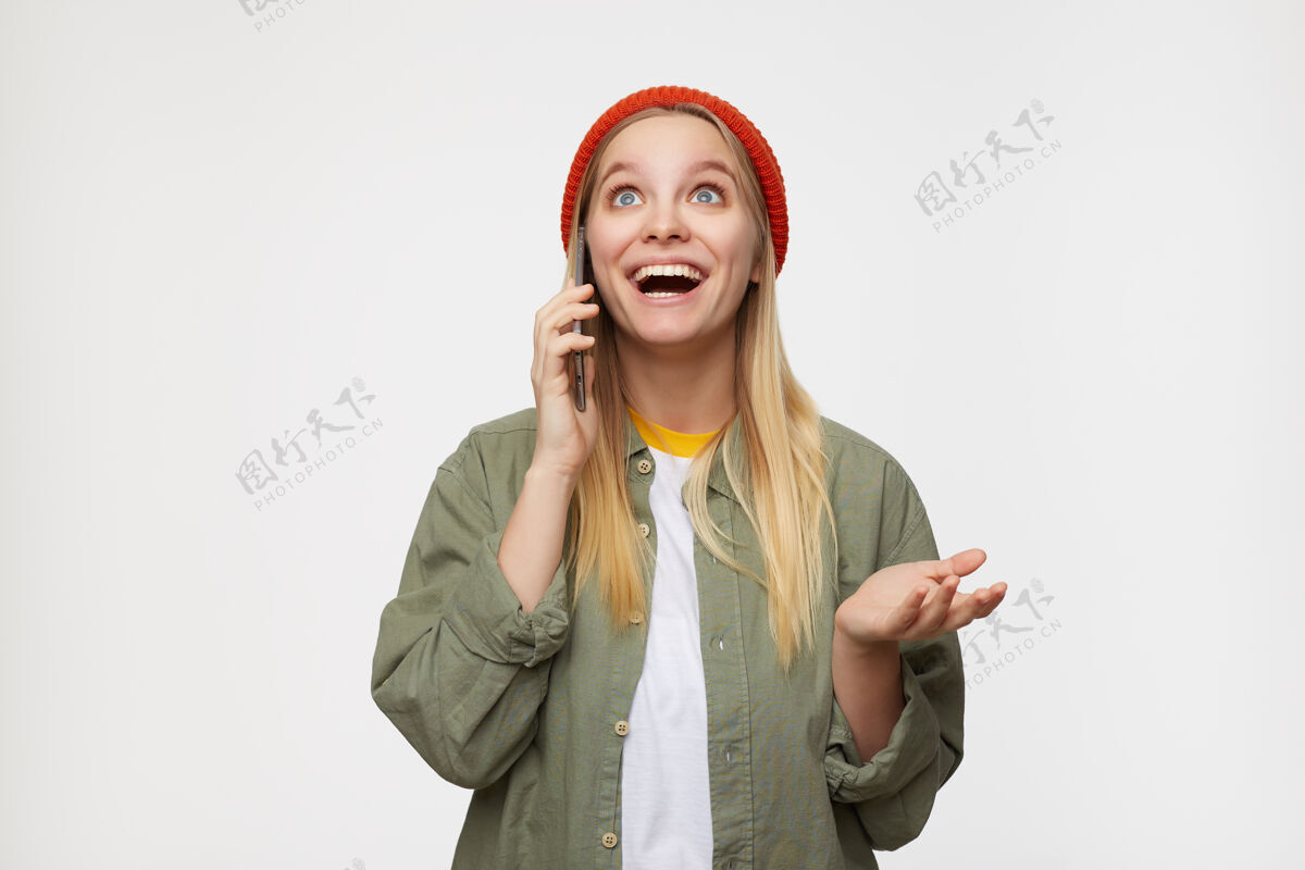 帽子惊讶的年轻蓝眼睛金发女性 随意的发型 愉快的电话交谈 兴奋地向上看 带着灿烂的微笑 孤立在蓝色手机表情蓝色