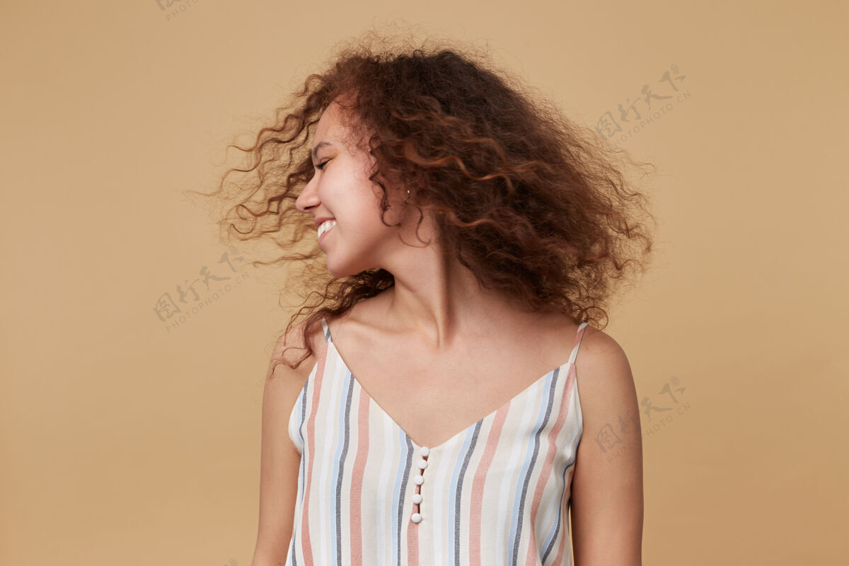 情绪年轻漂亮的棕发卷发女士 穿着夏装 站在米色的地板上 双手放在地上 挥舞着她的头 微笑着休闲穿着女性