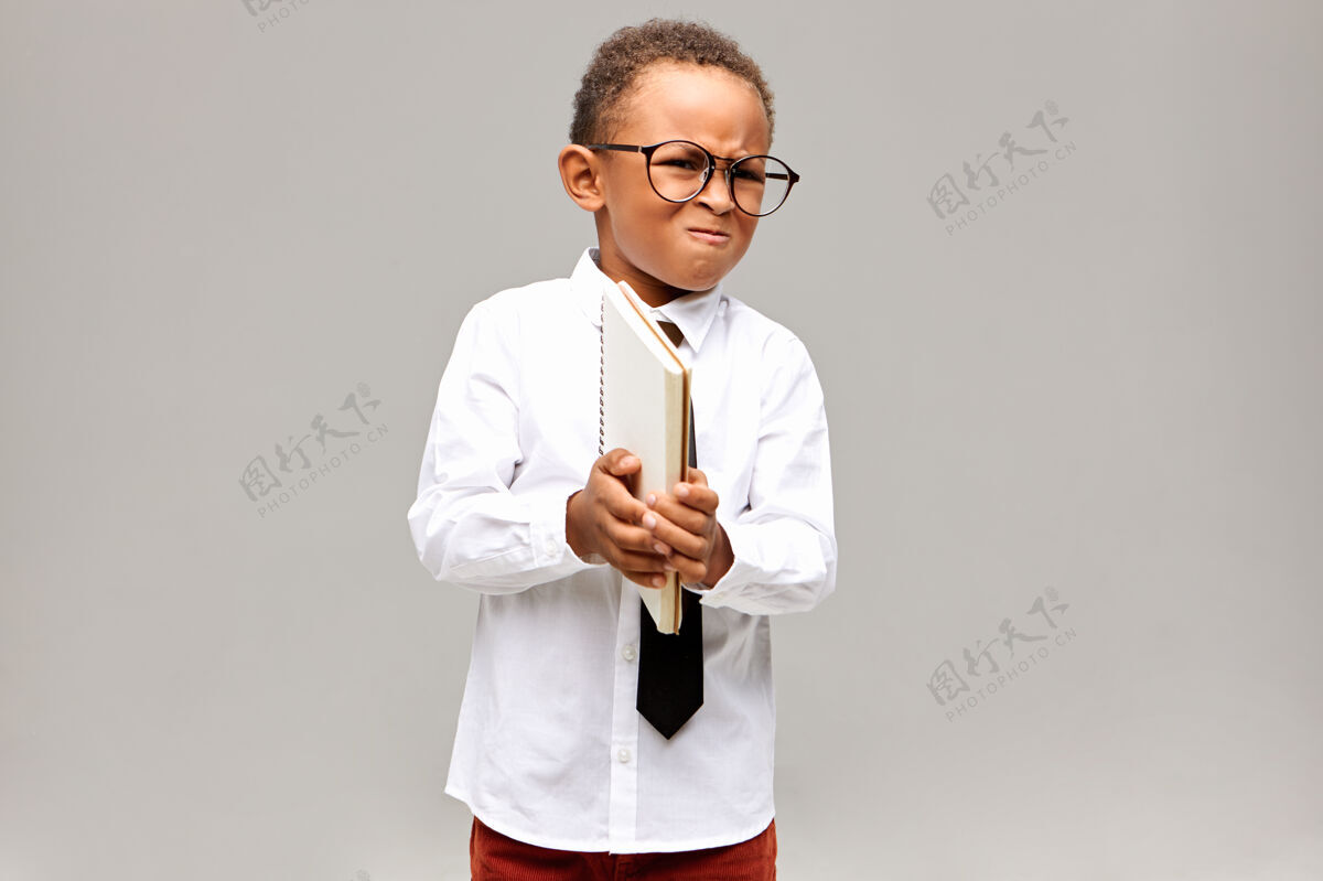 表情孩子 学习 教育和知识概念愤怒的非洲小男孩的画像 穿着白衬衫 戴着领带和眼镜 拿着抄写本 做鬼脸 因为他不会做数学而发疯情绪小学生站立